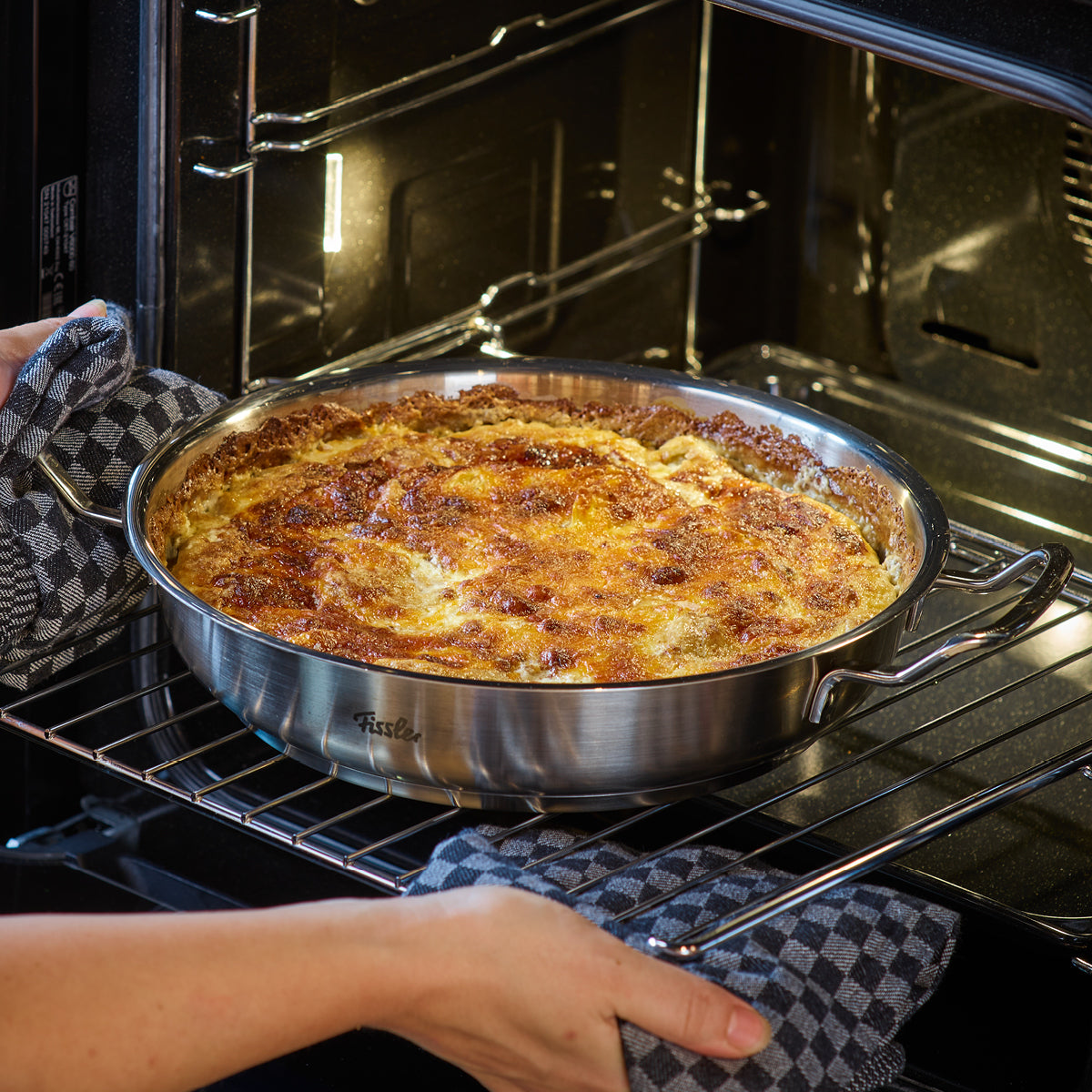 Eine Person schiebt mit karierten Handschuhen eine Fissler-Edelstahlpfanne mit goldenem, knusprigem Auflauf in den Ofen und zeigt damit deutsche Qualität.