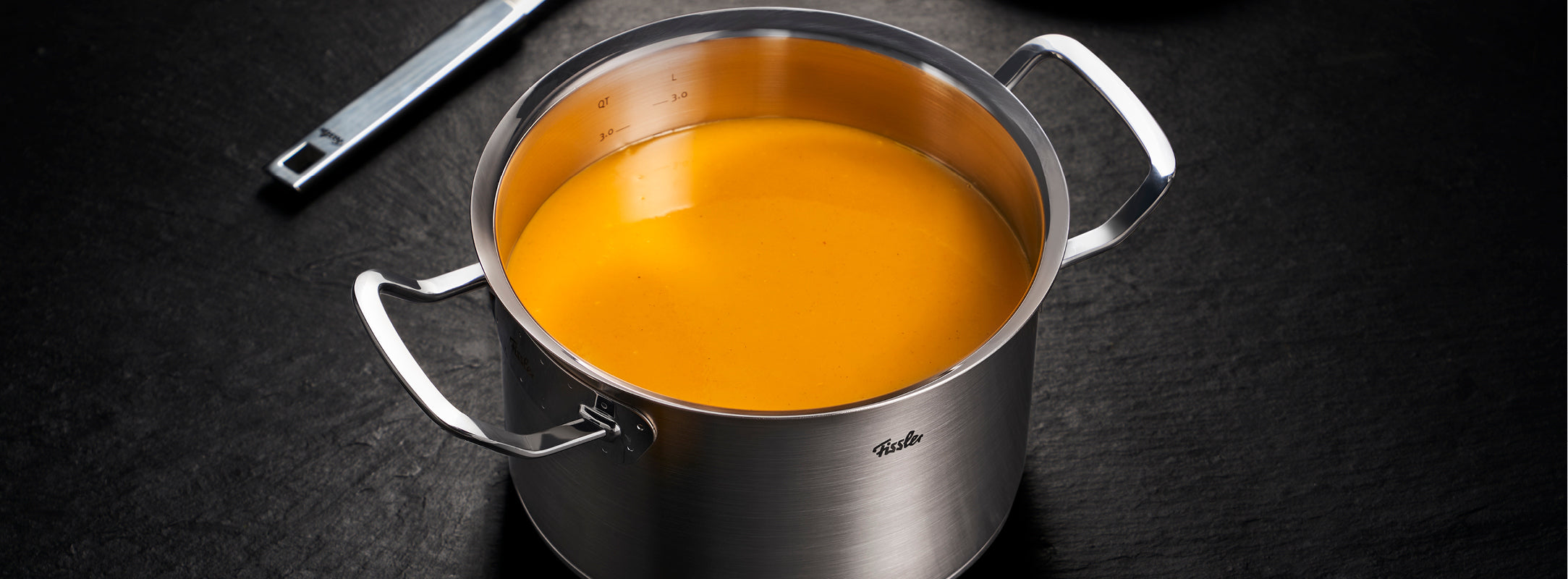Ein Fissler-Edelstahltopf mit orangefarbener Cremesuppe auf dunkler Oberfläche, begleitet von einer präzise gefertigten Schöpfkelle.