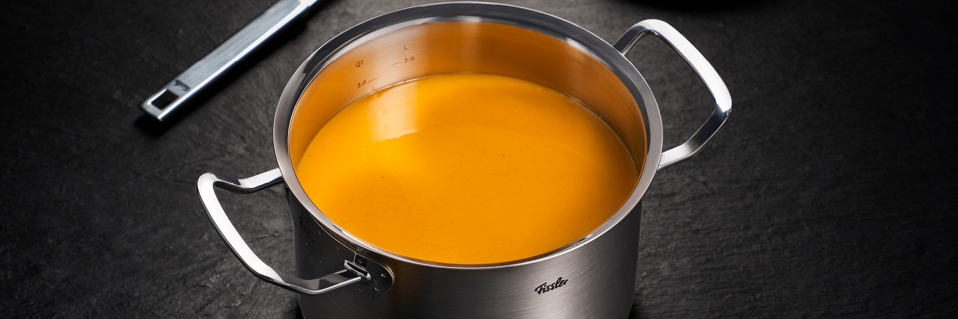 Ein in Deutschland präzisionsgefertigter Fissler-Edelstahltopf enthält eine orangefarbene Cremesuppe; dahinter befindet sich ein Schöpflöffelgriff aus Metall.