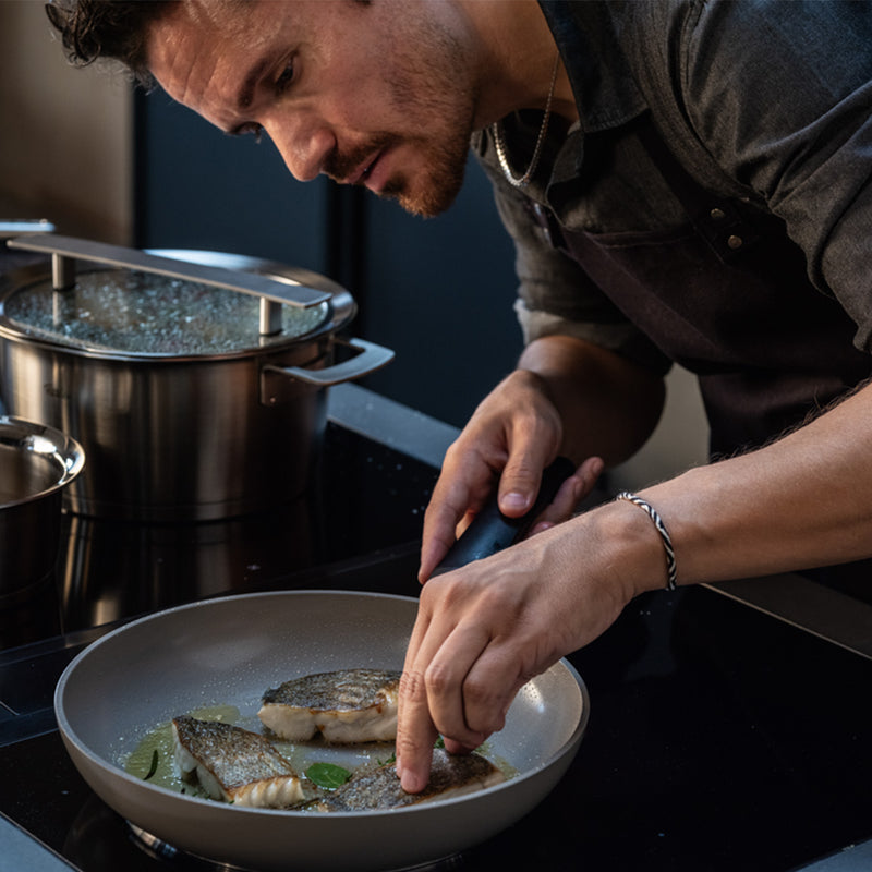 A man in a dark shirt and apron expertly prepares fish fillets in a Fissler Ceratal® Evo Classic Pfanne on the stovetop, finishing them with fresh herbs. The innovative nonstick ceramic surface of the pan delivers flawless results, while premium Fissler stainless steel pots—hallmarks of German craftsmanship and design—stand ready nearby.