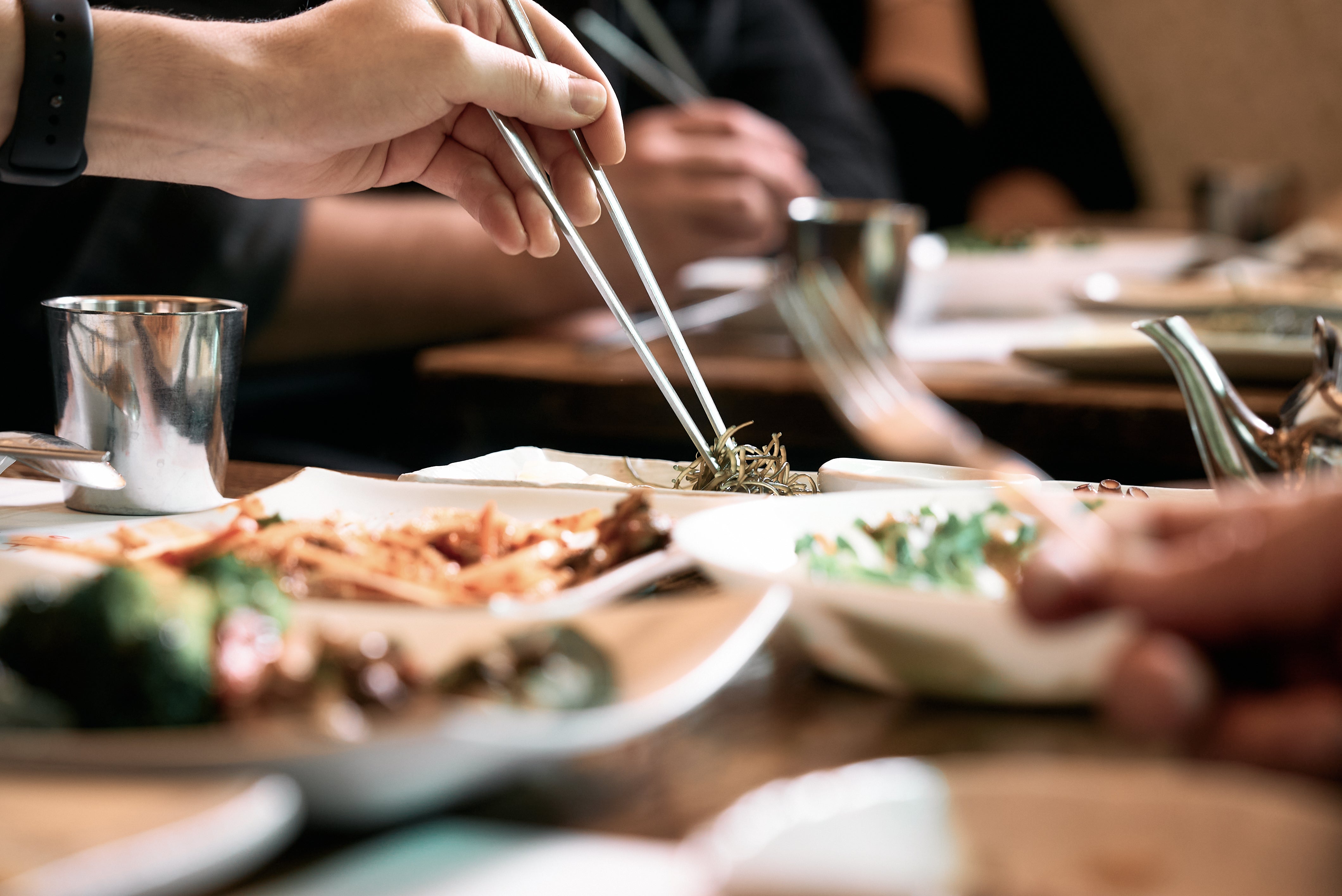 A person uses chopsticks to pick up food from a plate at a beautifully set table featuring various dishes served in elegant Fissler stainless steel cookware. The hands of others reach toward classic Fissler pots and pans, reflecting a shared meal that highlights precision engineering, lasting durability, and the timeless design of high-quality German-made kitchenware.