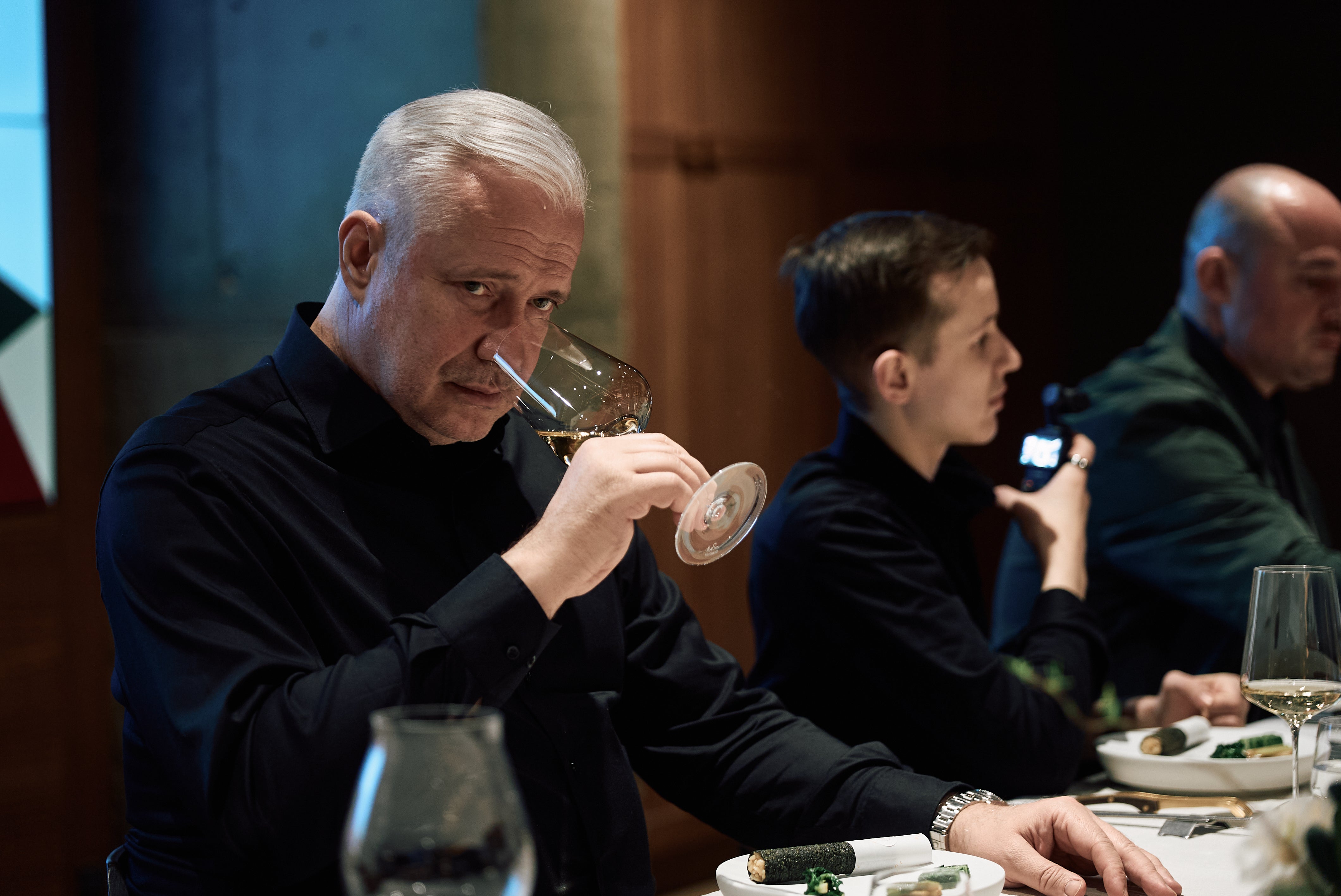 A man with gray hair in a dark shirt enjoys the aroma of a glass of wine at a dining table set with premium stainless steel Fissler cookware, reflecting precision engineering and timeless German design. Two other men share the moment nearby, one checking his smartphone, while the exceptional quality of Fissler pots and pans elevates their dining experience.