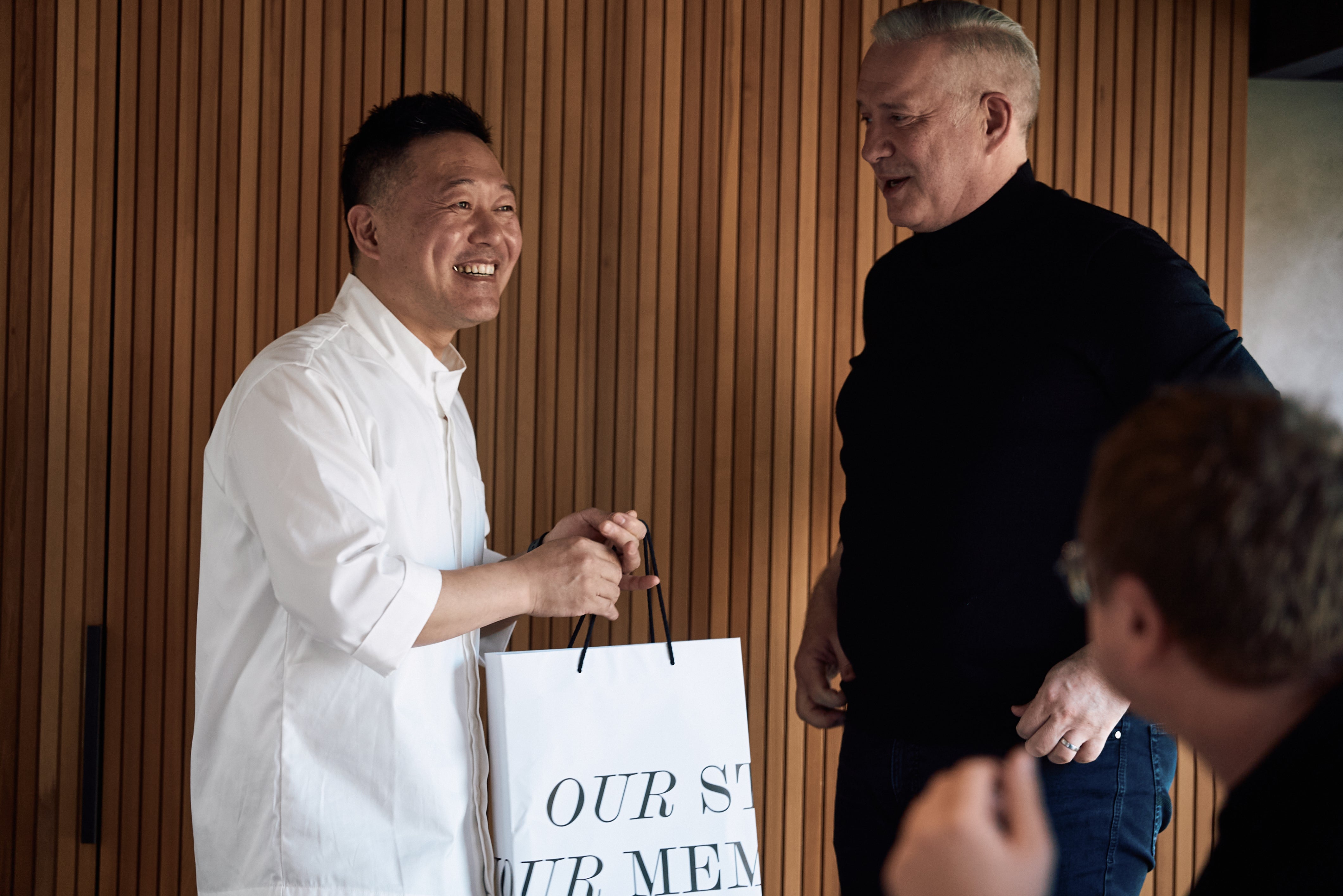 A smiling man in a white shirt holds a white Fissler-branded bag, standing beside another man in a black shirt. They are indoors with warm wood paneling behind them, highlighting the heritage and quality craftsmanship of Fissler's premium cookware products.