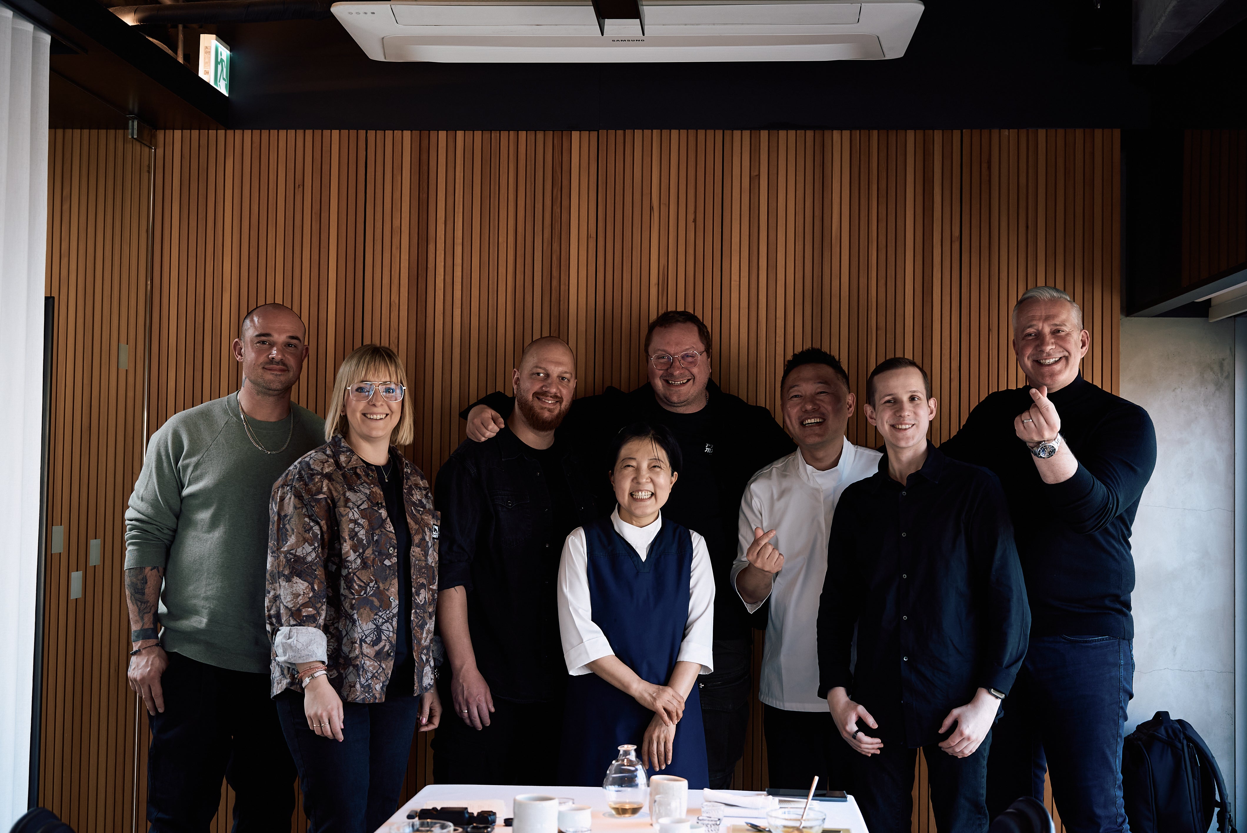 Eight people, representing the Fissler heritage of excellence since 1845, stand and smile together indoors in front of an elegant wood-paneled wall, exuding happiness and confidence. In the foreground, a white table displays high-quality Fissler cookware—stainless steel pots, pans, and kitchen accessories—embodying precision engineering, innovation, durability, and timeless design.