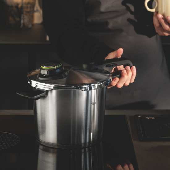A person holds a Fissler pressure cooker on a stovetop, another hand holds a mug. The kitchen background is softly blurred.