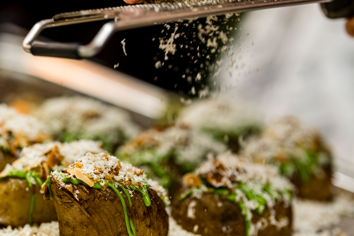 Close-up of a hand using a precision-engineered Fissler grater to add fresh cheese over golden roasted potatoes, elegantly garnished with vibrant green herbs and savory mushrooms. Fine cheese shavings fall delicately onto the dish, showcasing the quality and innovation of Fissler kitchen accessories.