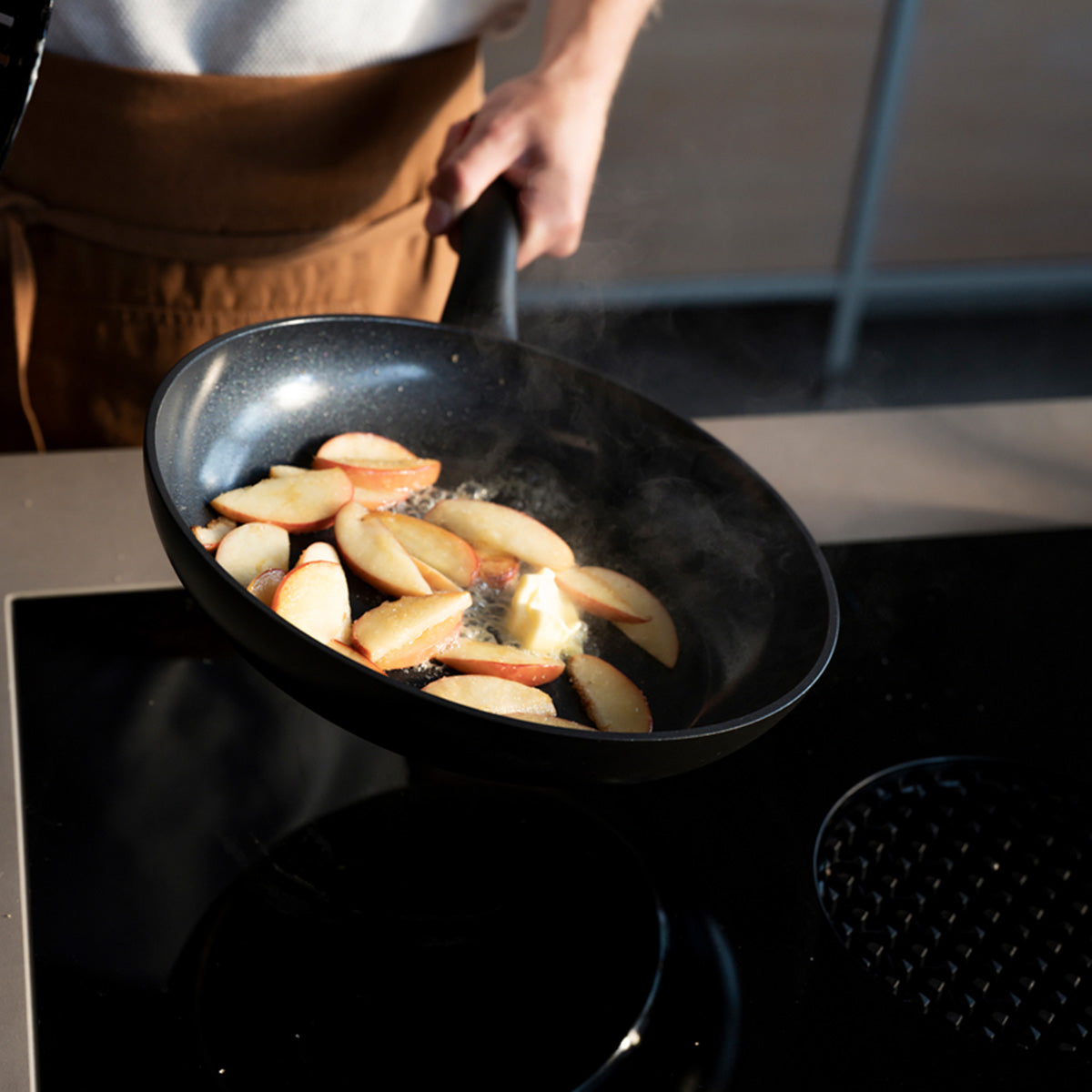 Sliced apples caramelizing in a Fissler Essential® pan set with rising steam, showing the chef's arm and torso representing precision Made in Germany.
