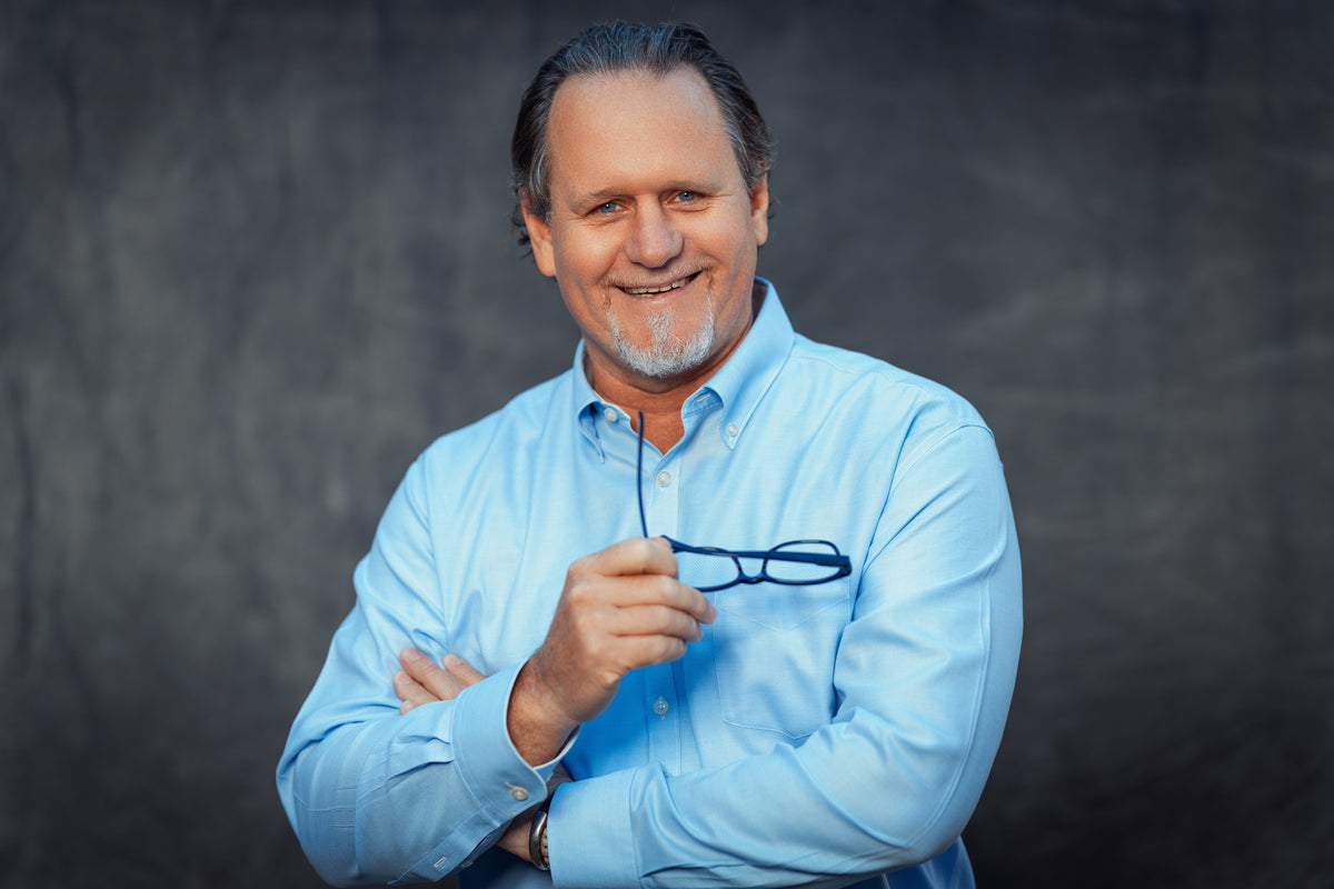 Distinguished middle-aged man with a gray beard and slicked-back hair, dressed in a light blue shirt, smiles warmly while holding eyeglasses in one hand. He stands poised against a dark, blurred background, reflecting Fissler’s heritage of confidence and timeless design—hallmarks shared by our precision-engineered stainless steel cookware proudly made in Germany.