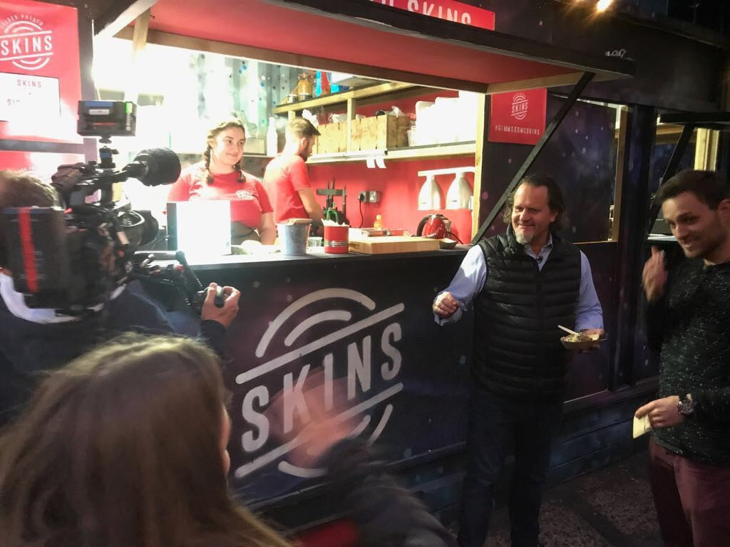A film crew captures the lively atmosphere at an outdoor food truck called SKINS at night, where two staff members in red shirts serve delicious meals through the window. Outside, two men interact and share a smile, showcasing the joy of good company and expertly prepared food. The vibrant scene reflects Fissler’s commitment to culinary excellence—rooted in German heritage since 1845—with high-quality stainless steel cookware ensuring every dish is made with precision, durability, and timeless design.