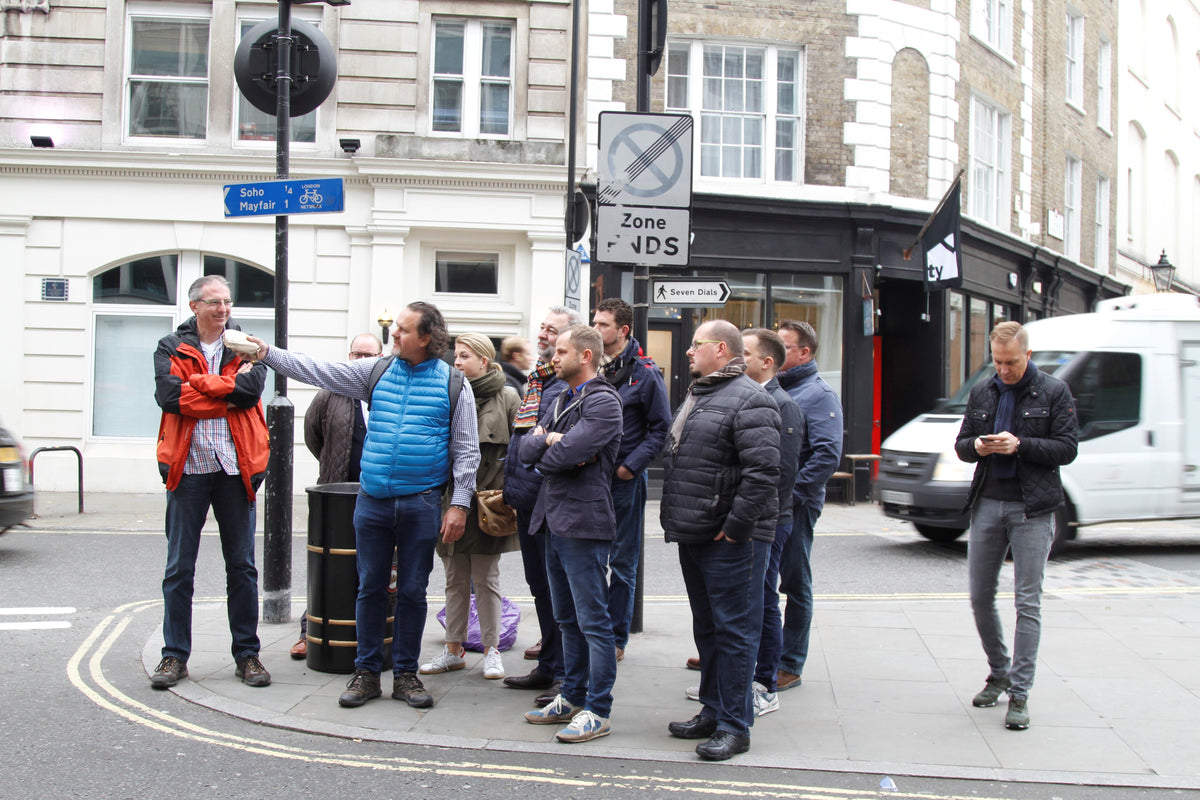 A group of people stand on a city sidewalk at a street corner, some engaged in conversation and others observing their surroundings with thoughtful attention, reflecting the spirit of connection and everyday life Fissler celebrates. One person checks their phone, highlighting modern living. Sturdy buildings and a passing van form the background—reminders of durable design and reliability, values mirrored in Fissler’s precision-engineered cookware crafted for timeless urban kitchens.