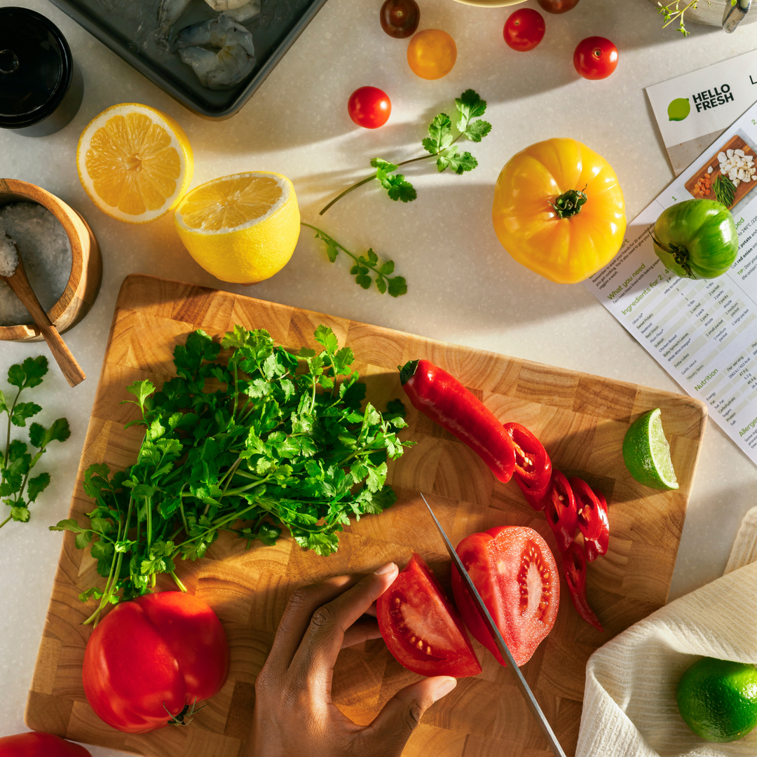 A hand expertly slices a ripe red tomato on a wooden cutting board, surrounded by fresh cilantro, red chili, lime, yellow tomato, lemon, and cherry tomatoes. The scene reflects meticulous preparation with precision kitchen tools reminiscent of Fissler’s German-engineered cookware and timeless culinary tradition.