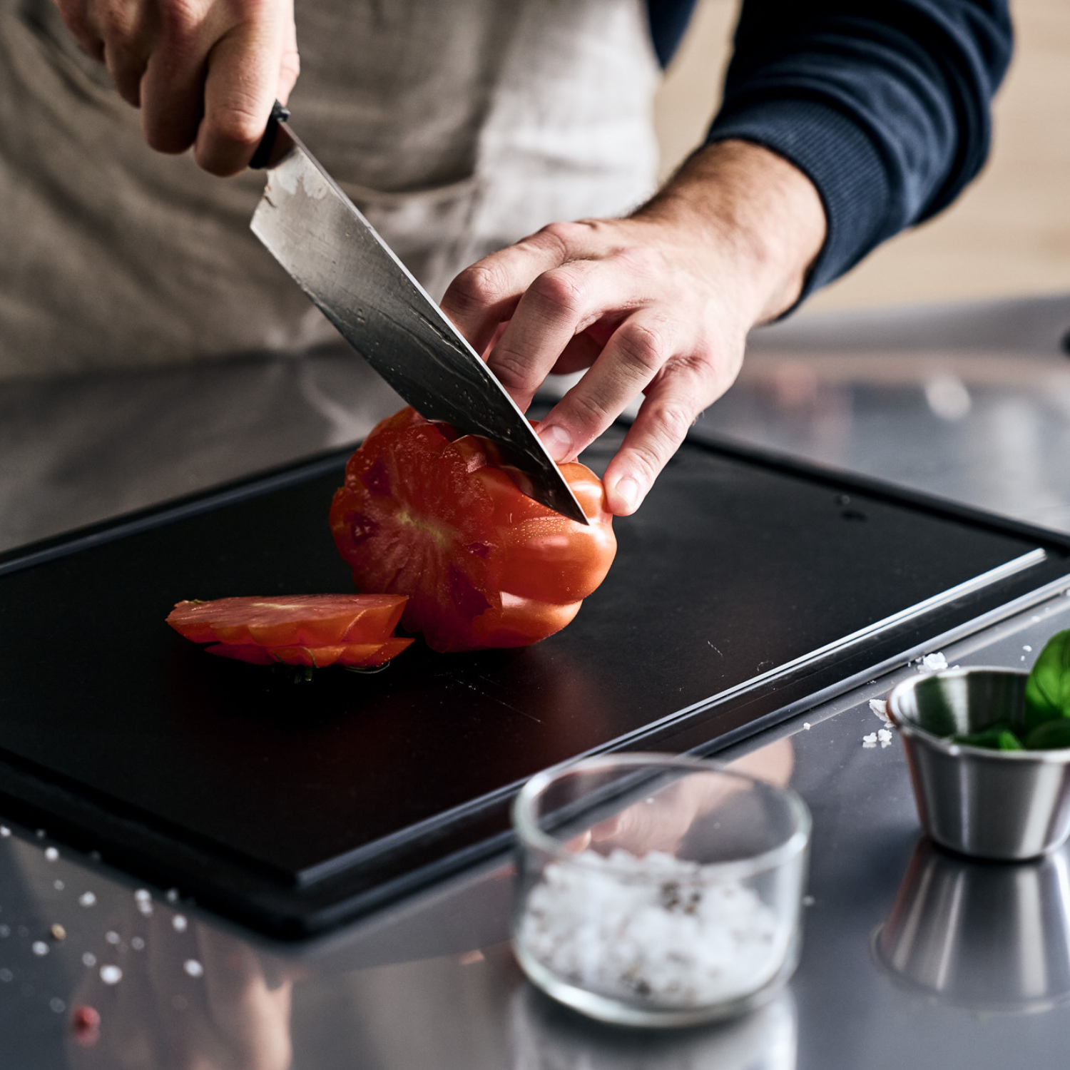 Ein Koch schneidet eine Tomate auf einem eleganten Fissler-Brett; grobes Salz, Basilikum und gestreute Körner liegen auf einer polierten Edelstahltheke.