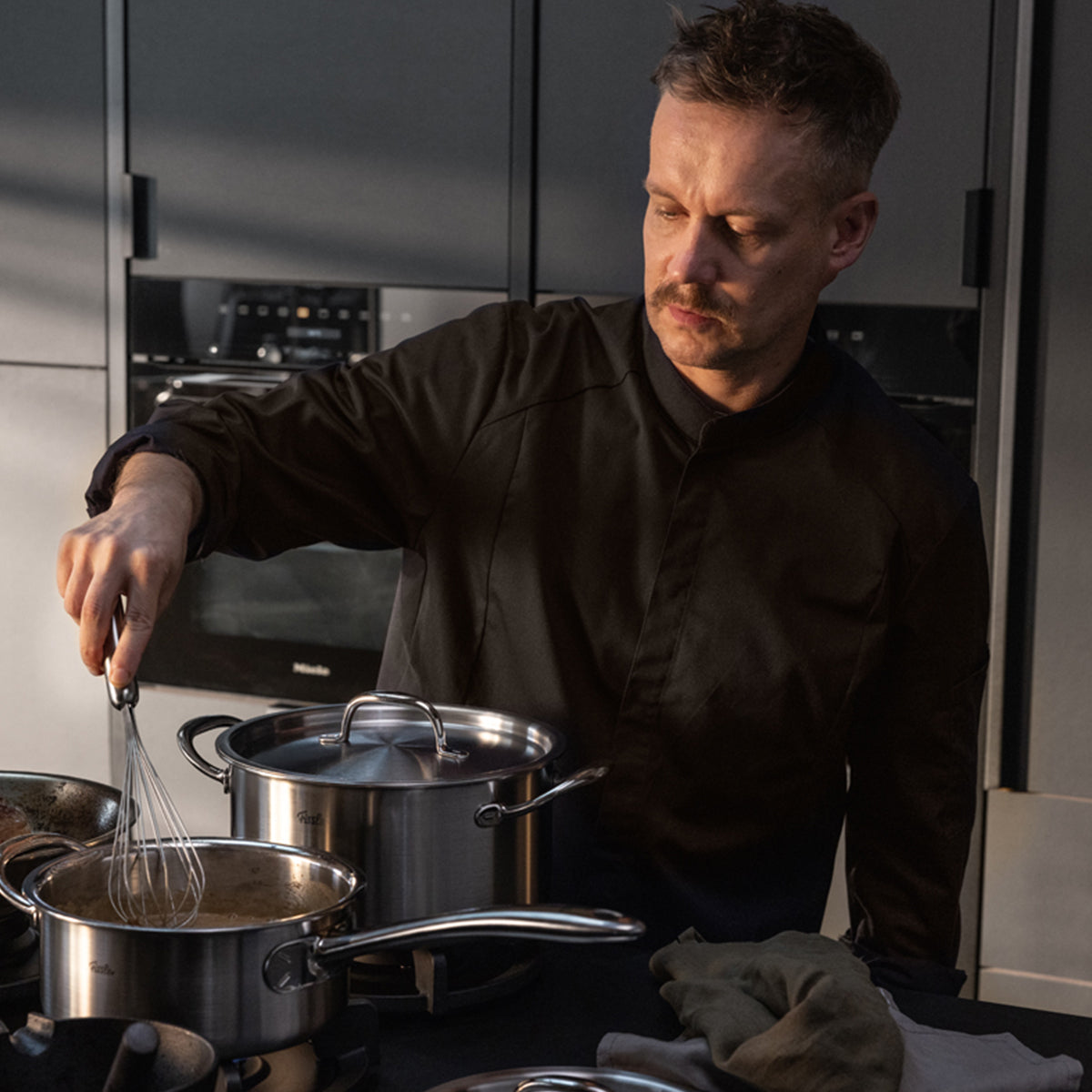 In a modern kitchen, a person in a dark chef's jacket skillfully stirs food with a whisk in the Fissler M5 Pro-Ply® Kochtopf mit Metalldeckel, expertly crafted from recycled stainless steel and engineered for optimal performance on an induction stove. Next to it sits another premium Fissler pot. Both high-quality pots embody Fissler's heritage of German precision, durability, and timeless design since 1845, and are dishwasher safe for effortless cleaning.