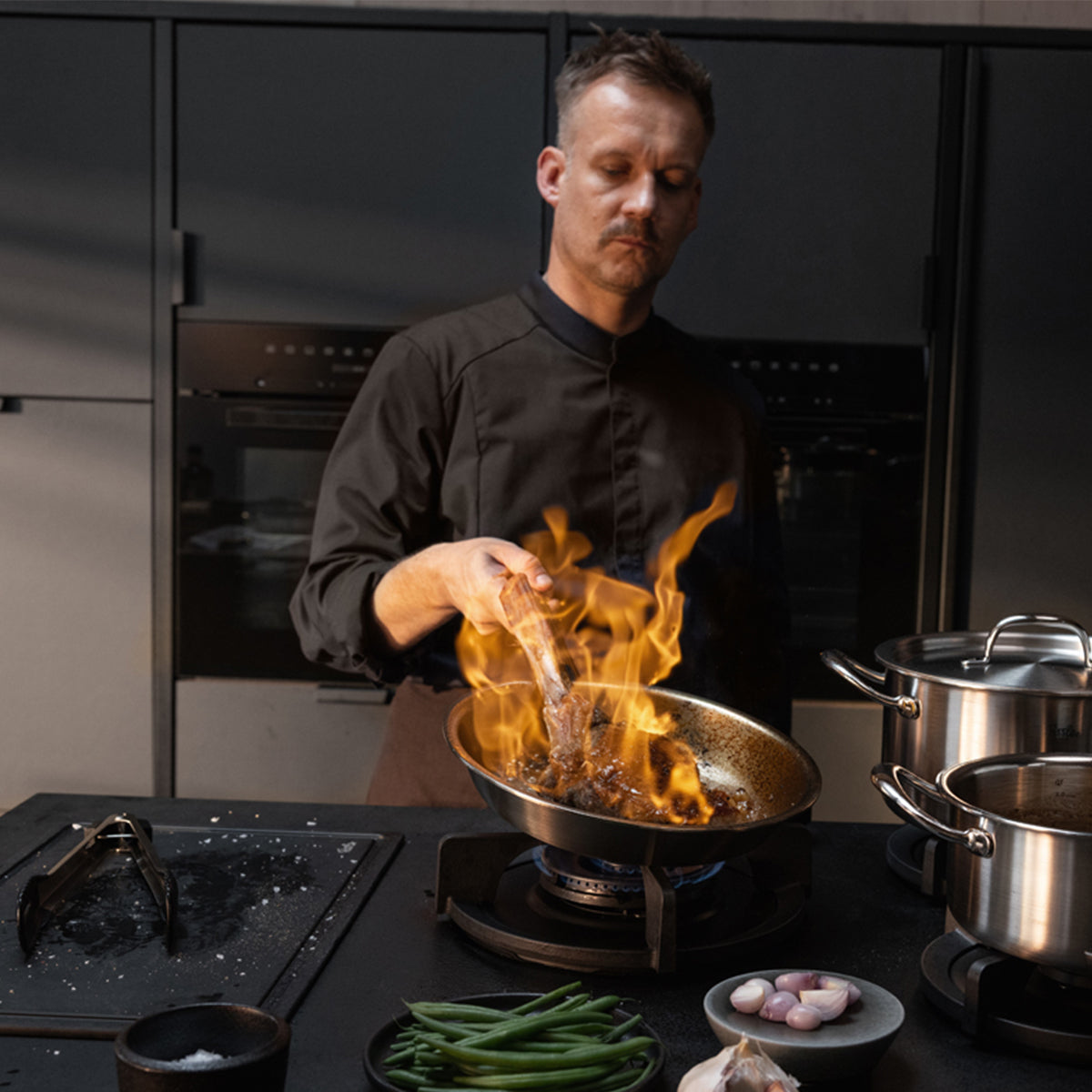 A chef in a black uniform expertly sears meat in the Fissler M5 Pro-Ply® pan made from recycled stainless steel, flames elegantly rising on a gas stove. On the countertop, high-quality Fissler stainless steel pots are arranged alongside fresh green beans, shallots, and salt, reflecting the brand’s heritage of precision engineering and timeless German design. All cookware shown is induction-ready and crafted for effortless cleaning—hallmarks of Fissler's innovation and durability since 1845.