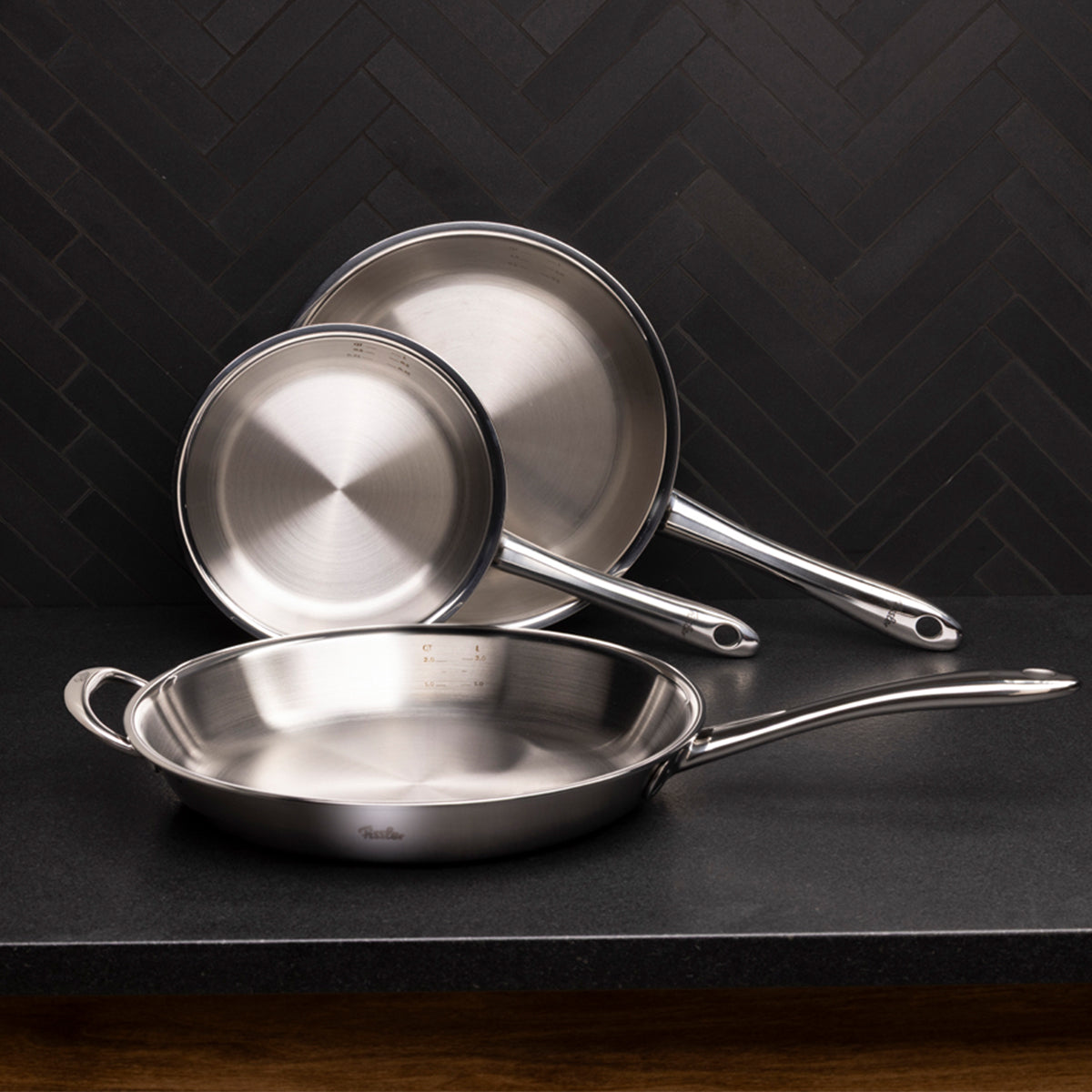 Drei Fissler M5 Pro-Ply® Pfannen aus hochwertigem, recyceltem Edelstahl in unterschiedlichen Größen sind sorgfältig gestapelt und auf einer dunklen Arbeitsplatte vor einer eleganten, schwarzen Küchenrückwand mit Fischgrätenmuster arrangiert. Die zeitlose, langlebige Qualität und das präzise Design von Fissler – Made in Germany seit 1845 – stehen im Mittelpunkt dieses stilvollen Küchenensembles.