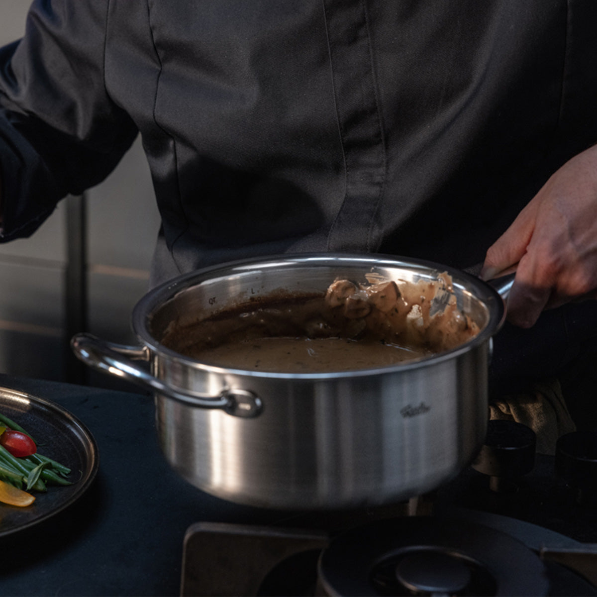 A person in a dark chef’s jacket expertly stirs a creamy brown sauce with a spatula in a Fissler M5 Pro-Ply® stainless steel pot with metal lid from the 3-piece set, positioned on the stove. The premium cookware, made in Germany and renowned for its precision engineering and timeless design, is suitable for both induction cooktops and ovens. Nearby, a plate of fresh vegetables highlights the versatility and quality that define Fissler’s heritage craftsmanship.