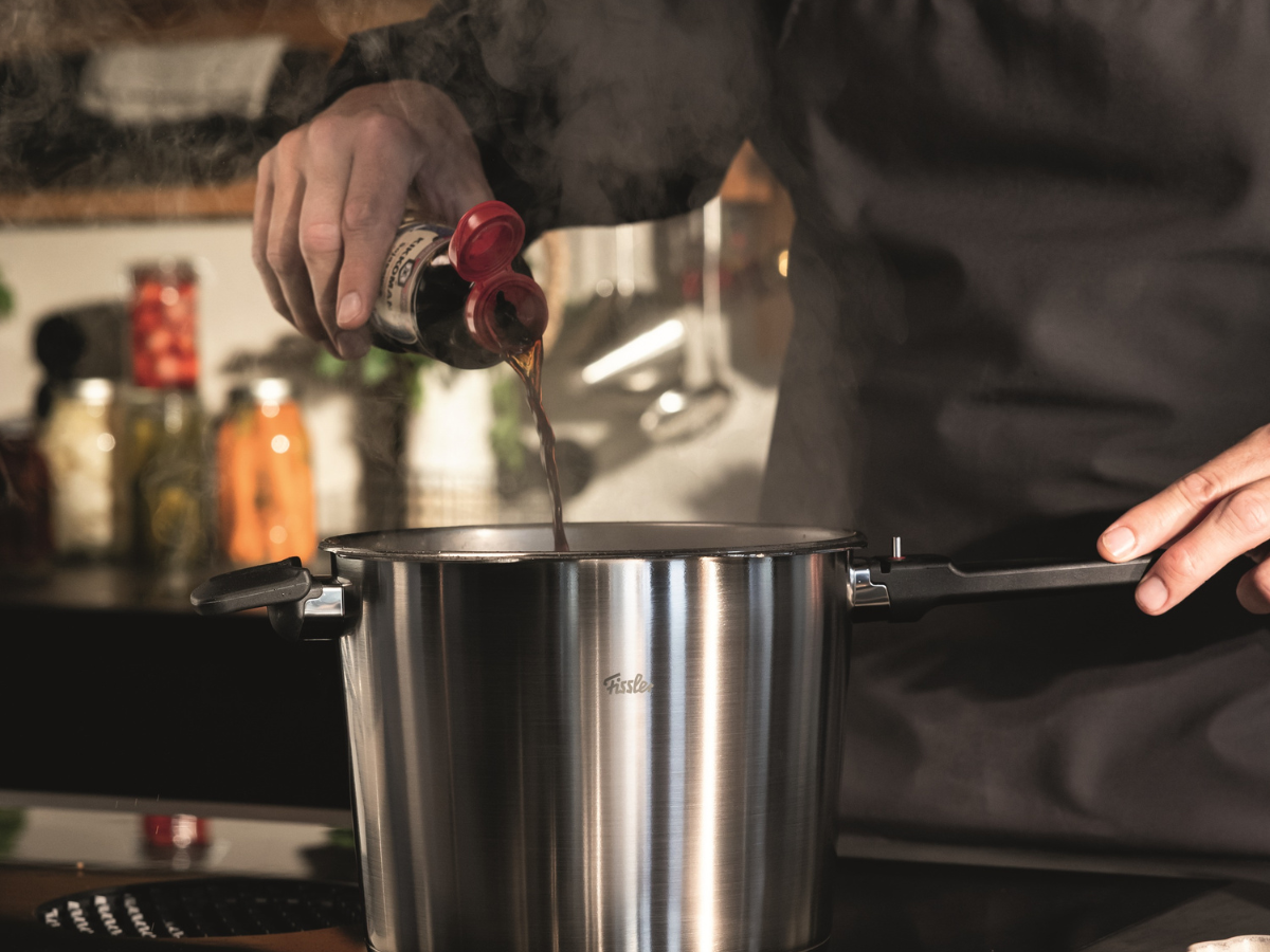 A person wearing a dark apron pours sauce from a bottle into a steaming Fissler stainless steel pot on a stovetop in a modern kitchen, showcasing the brand’s commitment to precision engineering, durability, and timeless German design.