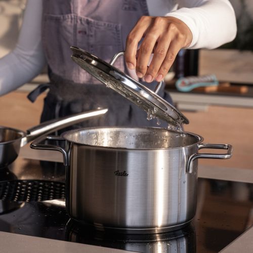 Eine Person hebt den Deckel eines Fissler-Topfes aus rostfreiem Stahl, der in Deutschland entwickelt wurde, während Dampf aufsteigt; ein anderer Fissler-Topf im Hintergrund.