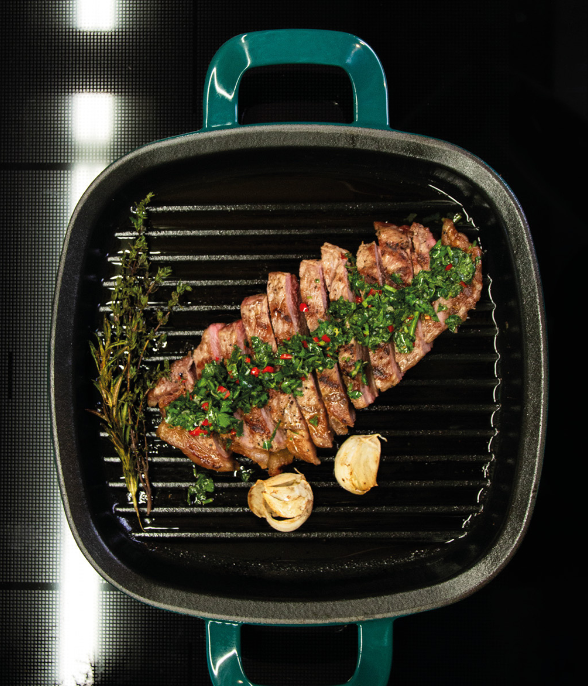 Juicy grilled steak with herb sauce, rosemary, and garlic in a black Fissler grill pan with characteristic green handles.