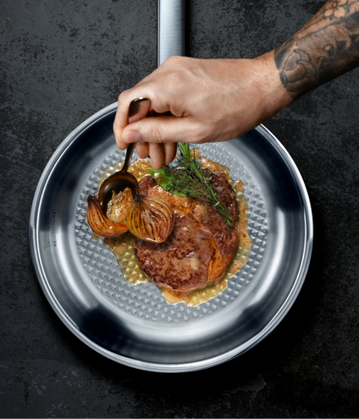 A tattooed hand spoons sauce over steak with grilled onions and herbs in a high-quality Fissler stainless steel pan on a dark surface.