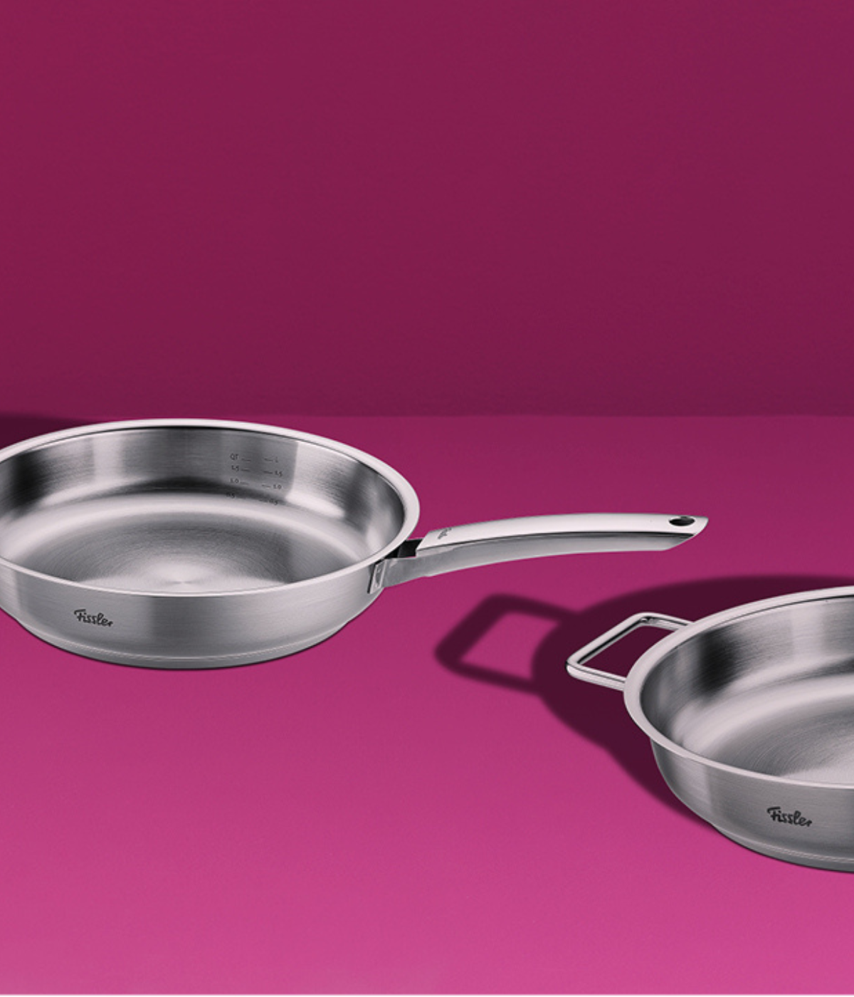 Fissler stainless steel frying pans - one with a long handle, one with a double handle - pictured on a pink surface with a matching background.