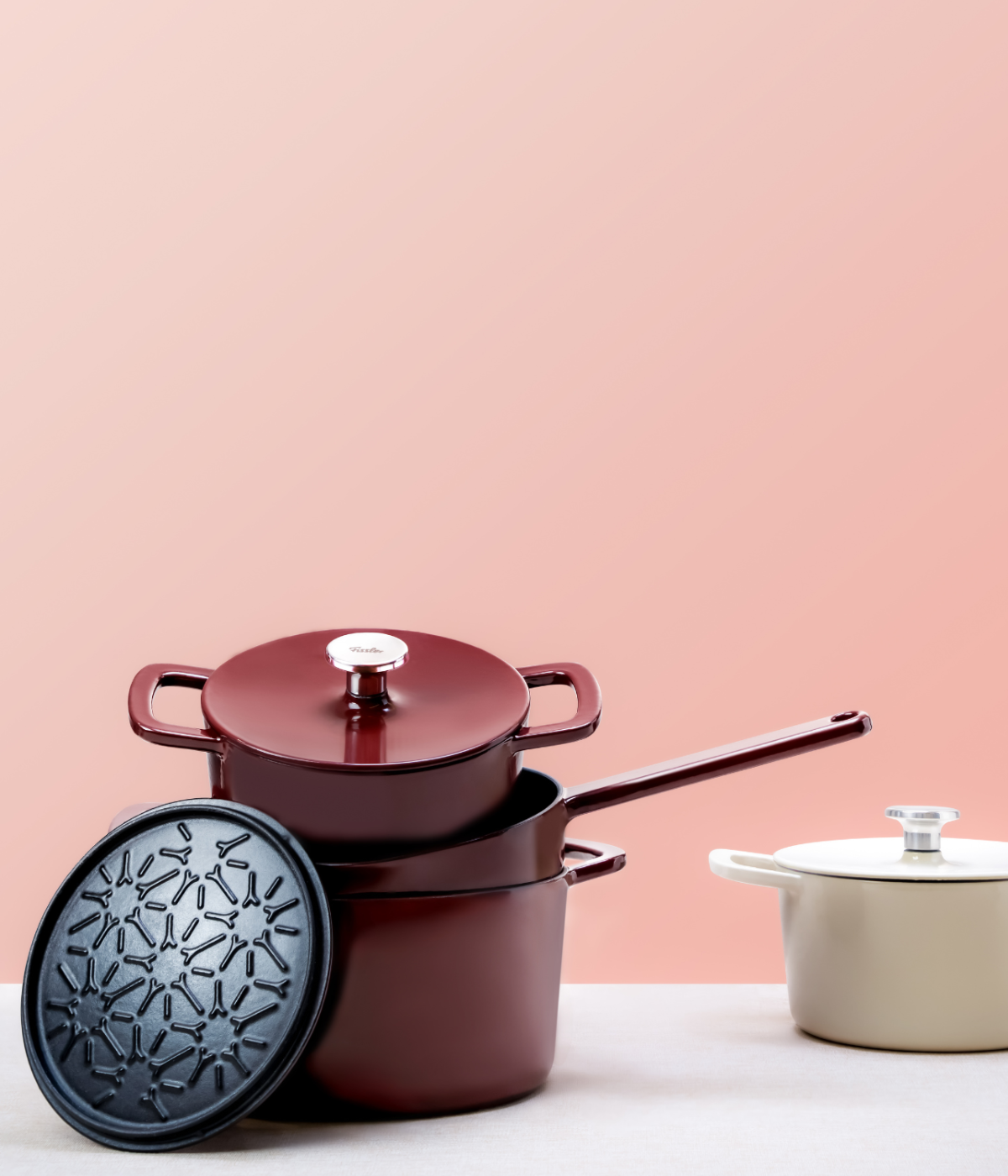 Fissler Cookware on a light surface with a pink background: chestnut and beige Dutch ovens, matching Cooking Pot and textured black Lid.