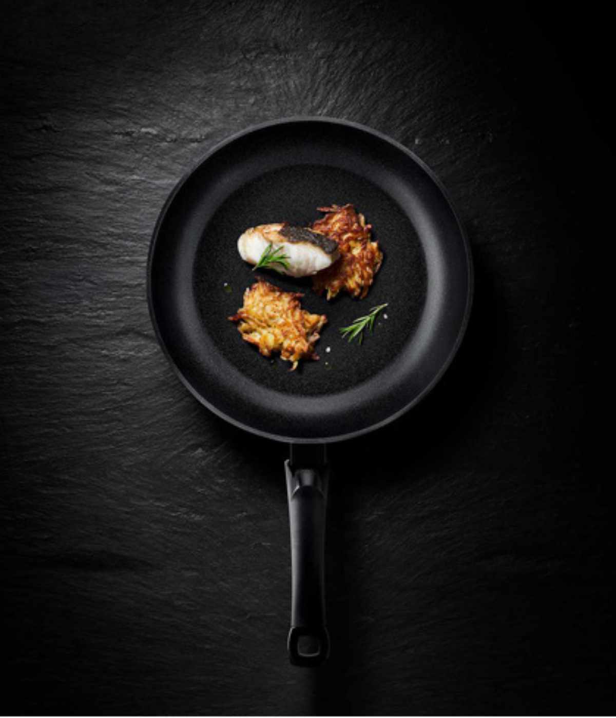 A black Fissler frying pan with cooked fish, fresh herbs, and two crispy roasted potatoes, reflecting German craftsmanship.