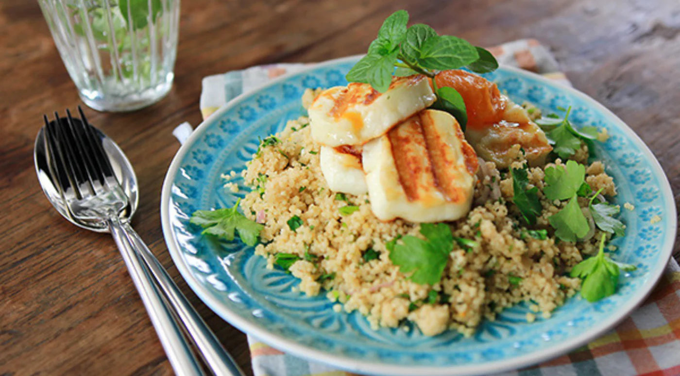 Auf einem Holztisch steht ein wunderschön angerichteter blauer Teller mit Couscous, gegrilltem Halloumi, frischen Kräutern und gerösteten Tomaten, dazu eine Gabel und ein Löffel – gefertigt in der bekannten Fissler-Edelstahlqualität – sowie ein Glas klares Wasser. Die elegante Anrichtung unterstreicht das präzise Design und den zeitlosen Stil, die Fisslers Tradition als Hersteller hochwertiger deutscher Kochgeschirrmarke widerspiegeln.