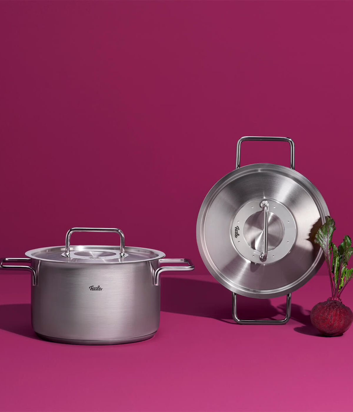 Fissler stainless steel pot and lid on a magenta surface, matching lid standing upright; beets with leaves add color.