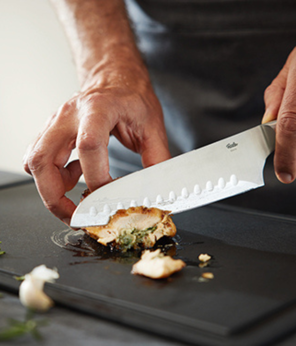 A person expertly slices stuffed chicken breast on an elegant Fissler cutting board with a precision chef's knife, with herbs and garlic nearby.
