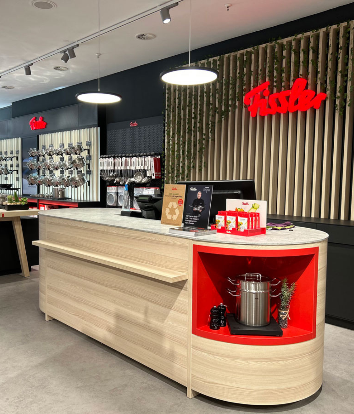 A Fissler stainless steel pot on an elegant wooden counter, with high-quality cookware and the illuminated Fissler logo in the background.