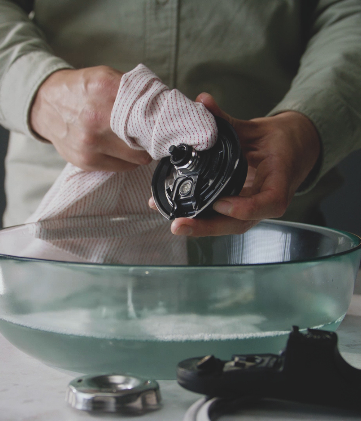 A person cleans the blade of a Fissler blender over a glass bowl of water; other high-quality blender parts lie on the counter next to it.