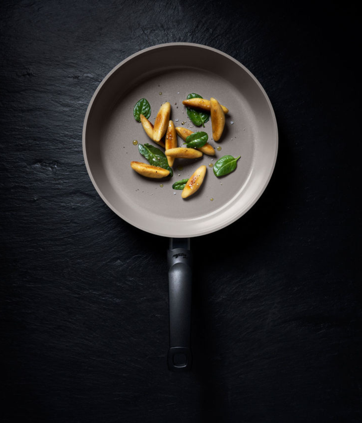 Fissler non-stick frying pan in beige with black handle, showing golden gnocchi and basil leaves on a dark slate background.