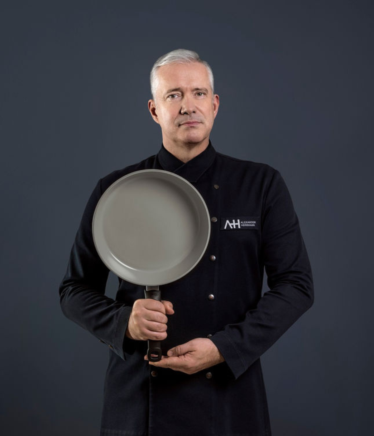 A man with short gray hair in a dark chef's jacket presents a Fissler frying pan upright against a simple, dark background.