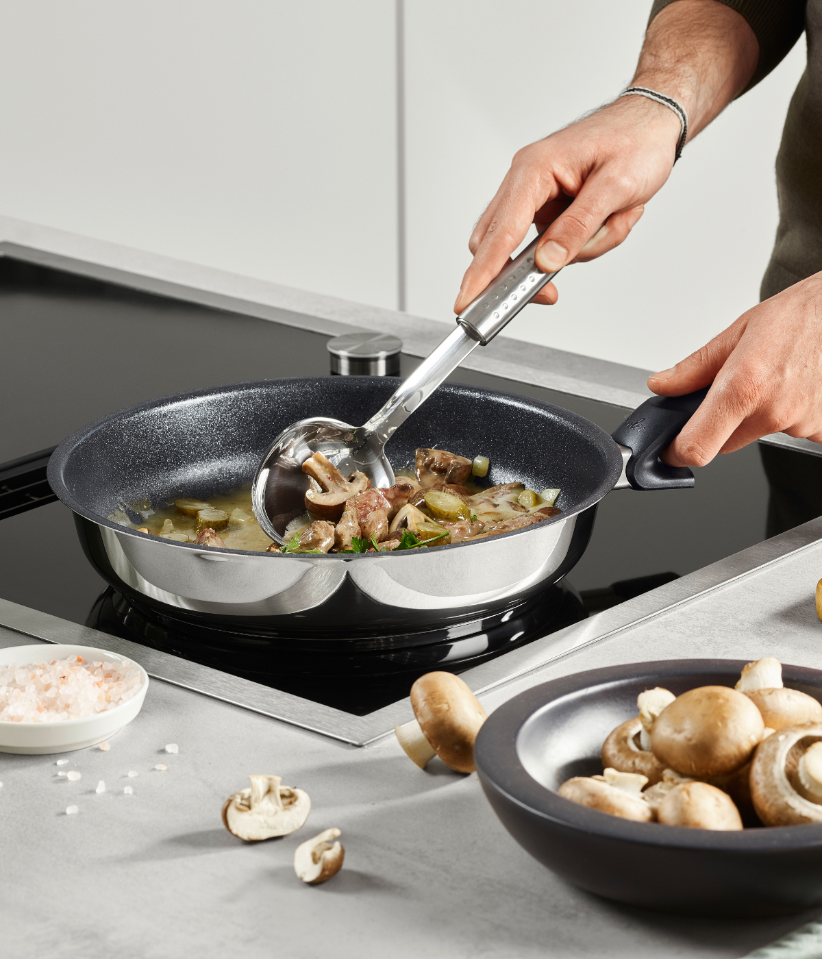 A person stirs meat and vegetables in a Fissler stainless steel pan; mushrooms and a bowl of pink salt sit on the counter nearby.