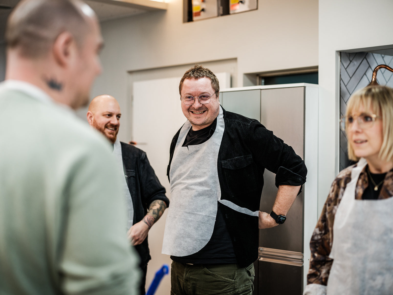 Three adults gather in a modern kitchen, smiling and engaging in conversation. The man at the center, wearing an apron and glasses, stands beside gleaming Fissler stainless steel cookware on the countertop. To his right, a woman in glasses listens attentively, while another man appears partially on the left. Premium German-engineered pots and pans add a touch of timeless design and durability to their shared cooking experience.