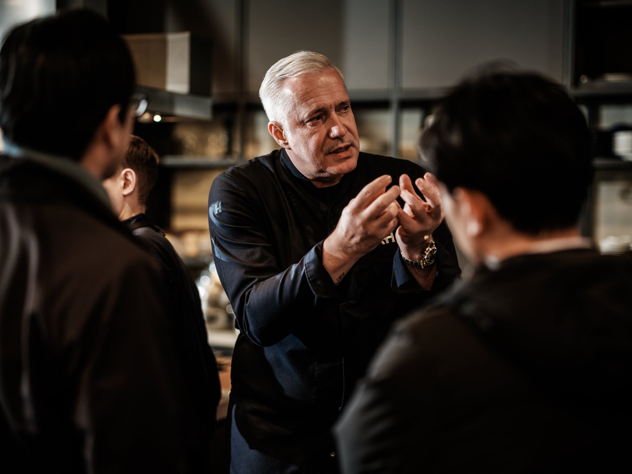 A distinguished chef with gray hair passionately explains a cooking technique to three attentive listeners in a modern kitchen, gesturing expressively above premium Fissler stainless steel pots and pans. The scene highlights the chef’s expertise alongside Fissler’s commitment to precision engineering, innovation, and timeless German craftsmanship.
