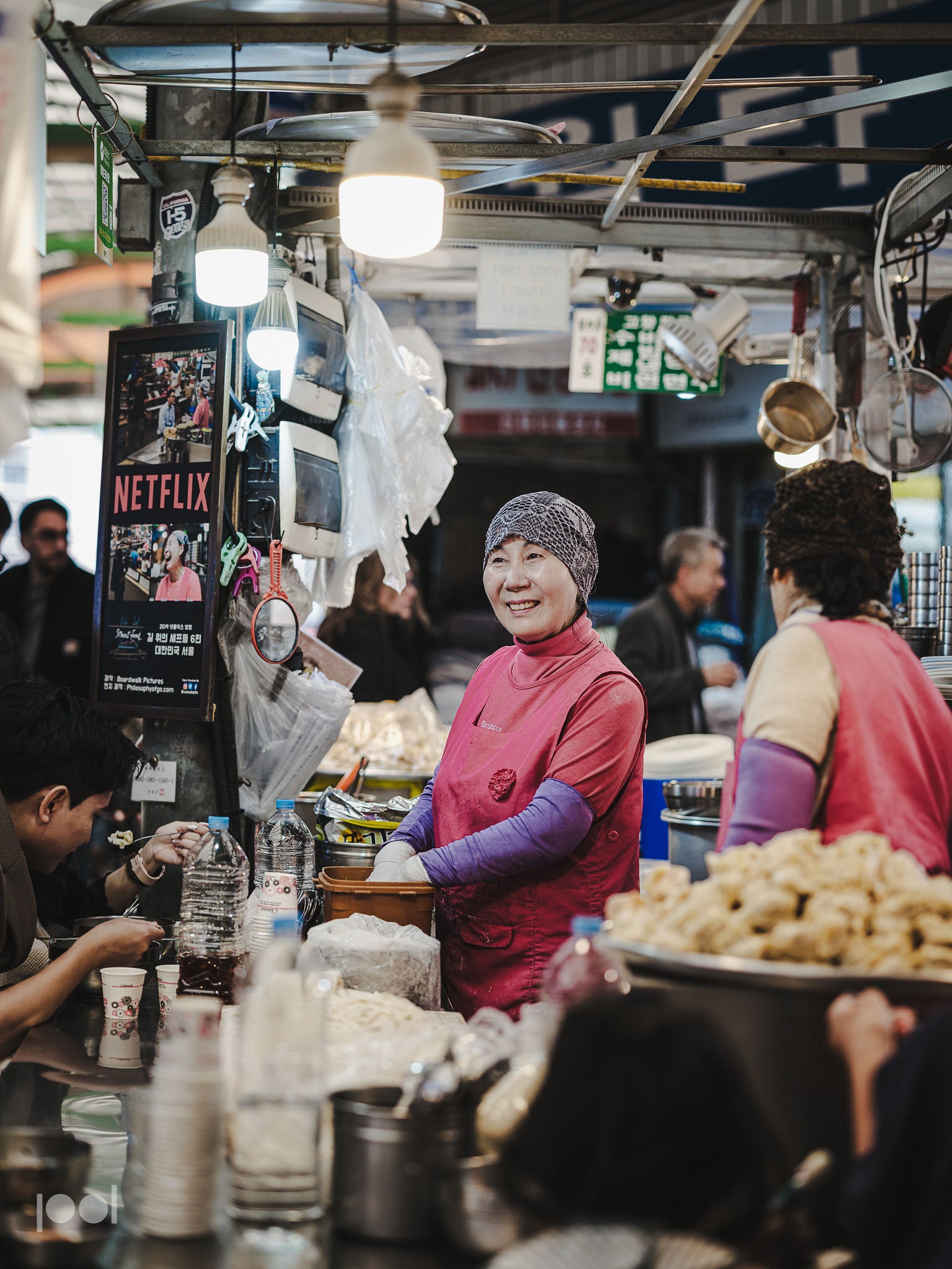 A smiling woman in a red apron skillfully prepares food at a bustling market stall, surrounded by customers, fresh ingredients, and warm hanging lights. She uses premium stainless steel cookware and utensils, reflecting Fissler’s tradition of precision engineering and timeless German design. A Netflix poster is visible in the background.