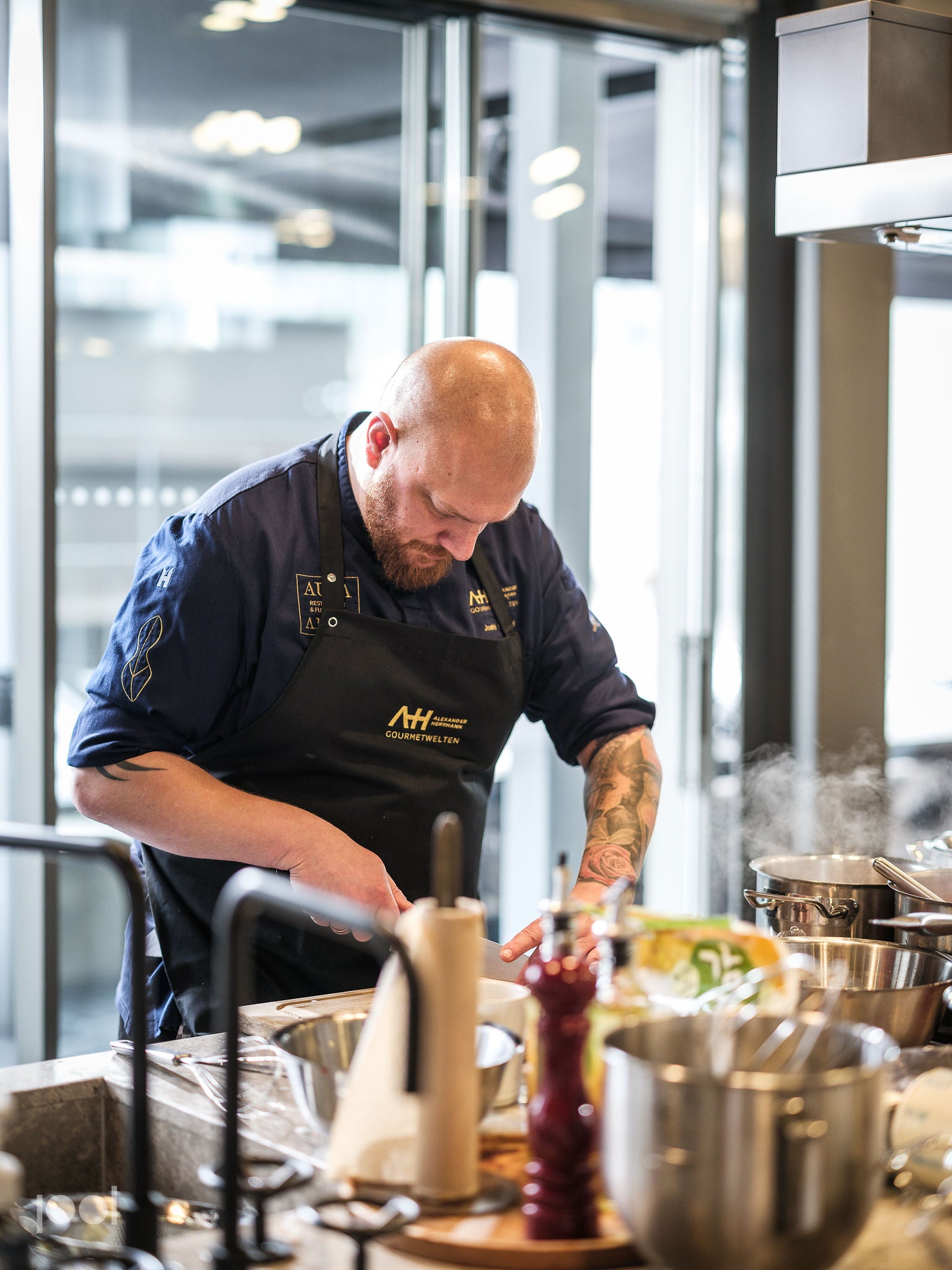 A chef in a navy apron expertly chops ingredients at a modern kitchen counter, surrounded by premium Fissler stainless steel pots, pans, and accessories. Sleek bowls and utensils highlight the timeless design and precision engineering of Fissler cookware, while steam rises from the durable pots—showcasing innovation and German craftsmanship since 1845.