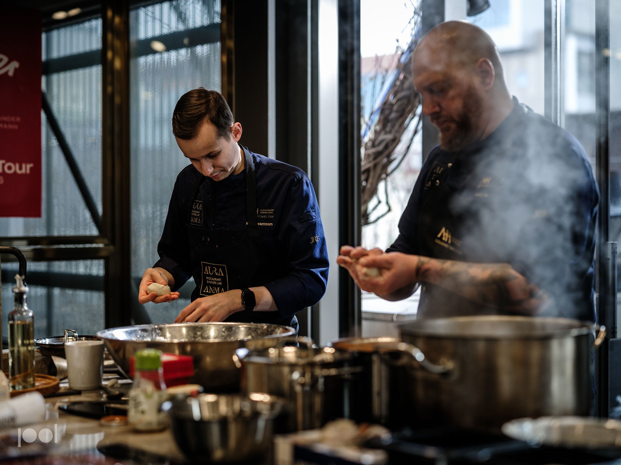 In a sunlit kitchen, two chefs work together surrounded by Fissler stainless steel cookware. One chef expertly shapes dough at the counter beside large mixing bowls, while the other tends to steaming Fissler pots and pans on the stove. Broad windows behind them fill the scene with natural light, highlighting the precision engineering and timeless design of Fissler products in use.